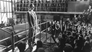 There is a picture of a man standing on a platform, addressing his workers. It is a scene from the movie, Schindler's List. It is one of the best movies based on books. 